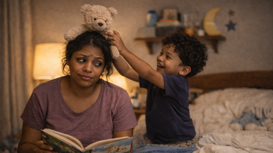 Image of a small boy playfully putting his teddy bear on his mum's head as they are both sat on the bed at night. The mum looks tired as she is trying to read a calming bedtime story.